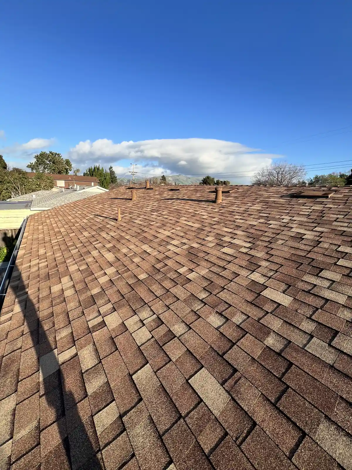 New brown blend shingle roof with two stone-coated pipe sleeves, gutter edge at left, white clouds above