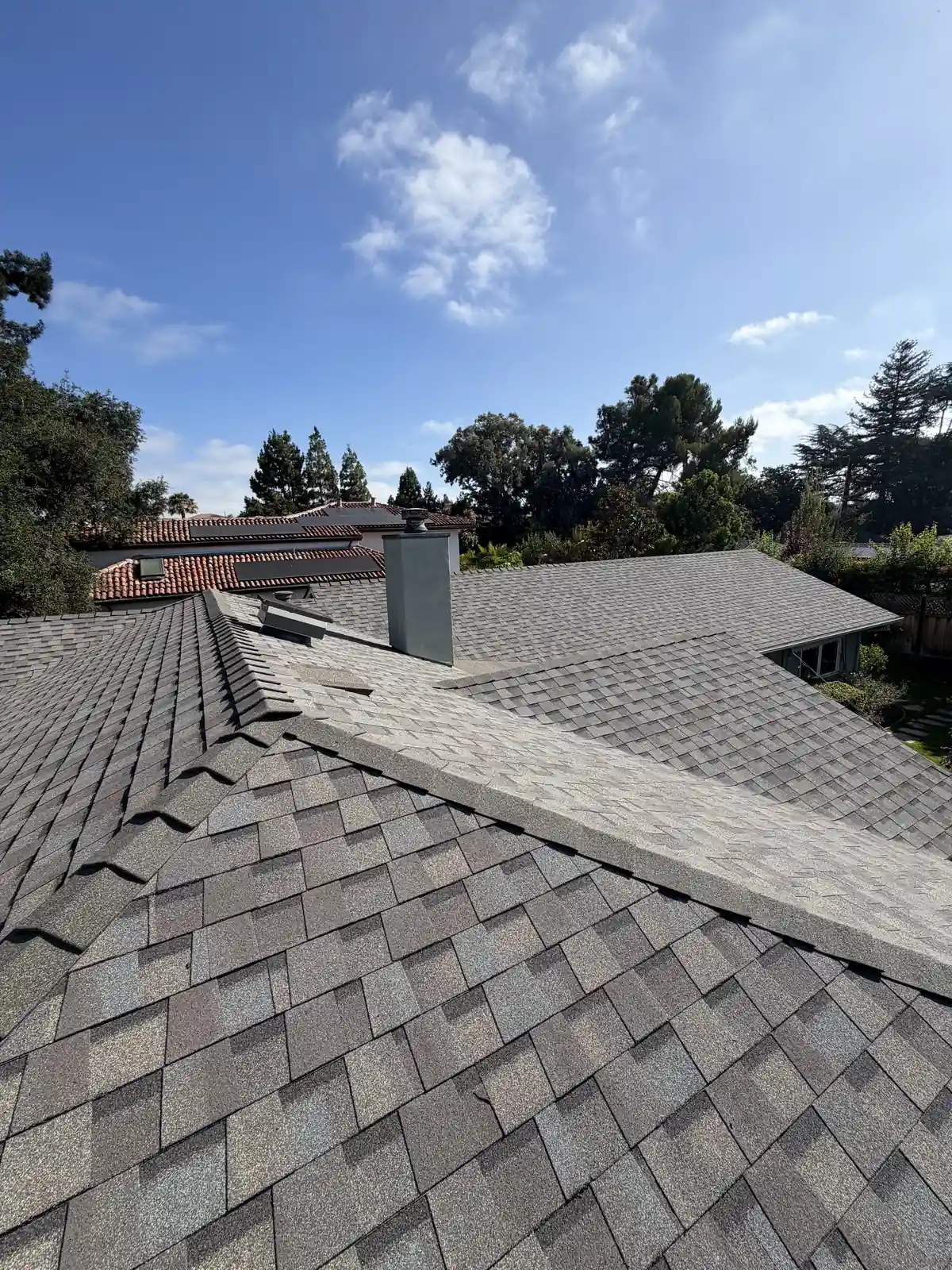 New gray shingle roof on a house with a stucco chimney, a cloud in the blue sky, oaks around